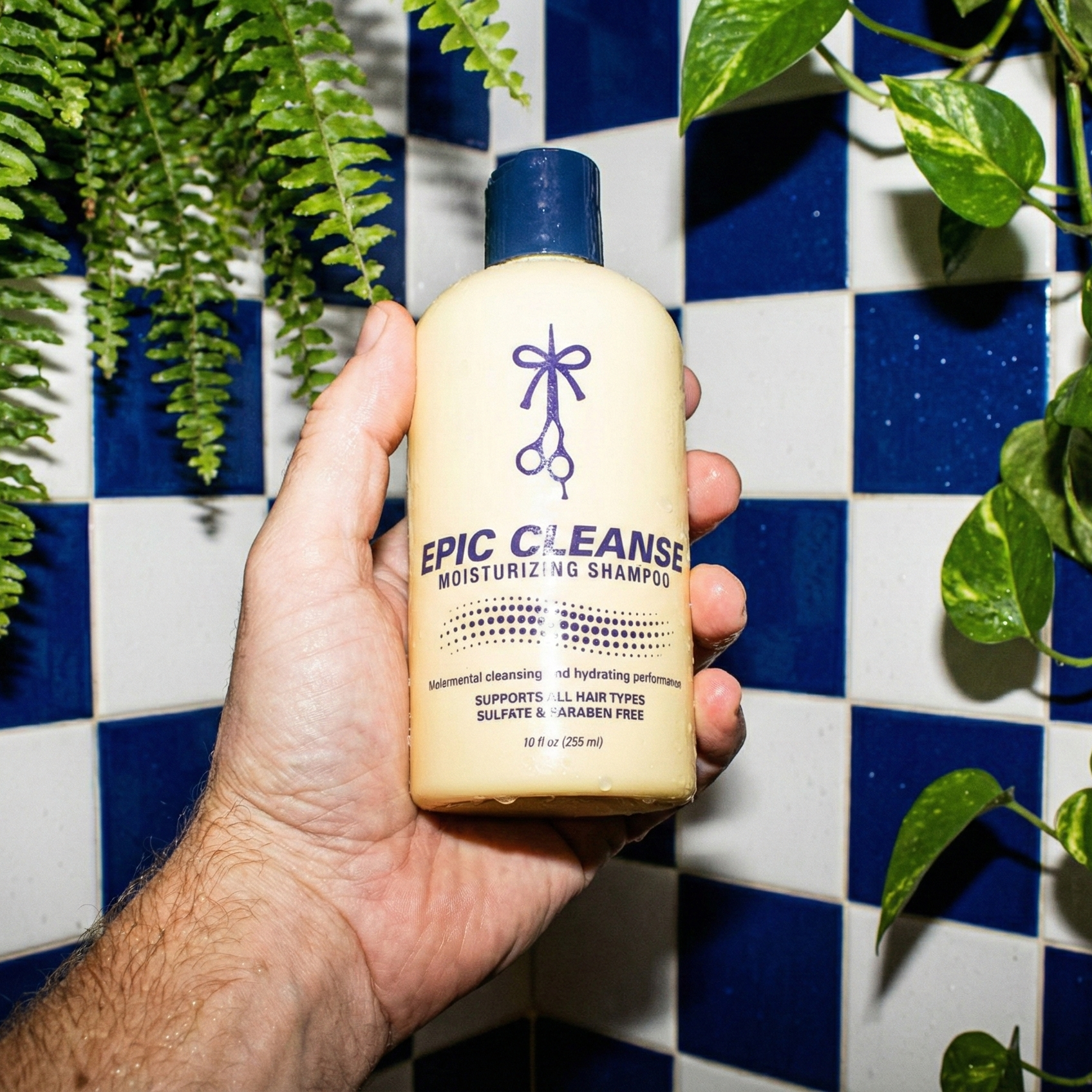 Hand holding a bottle of 'Epic Cleanse' shampoo against a blue and white checkered wall with plants.