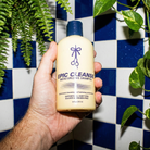 Hand holding a bottle of 'Epic Cleanse' shampoo against a blue and white checkered wall with plants.